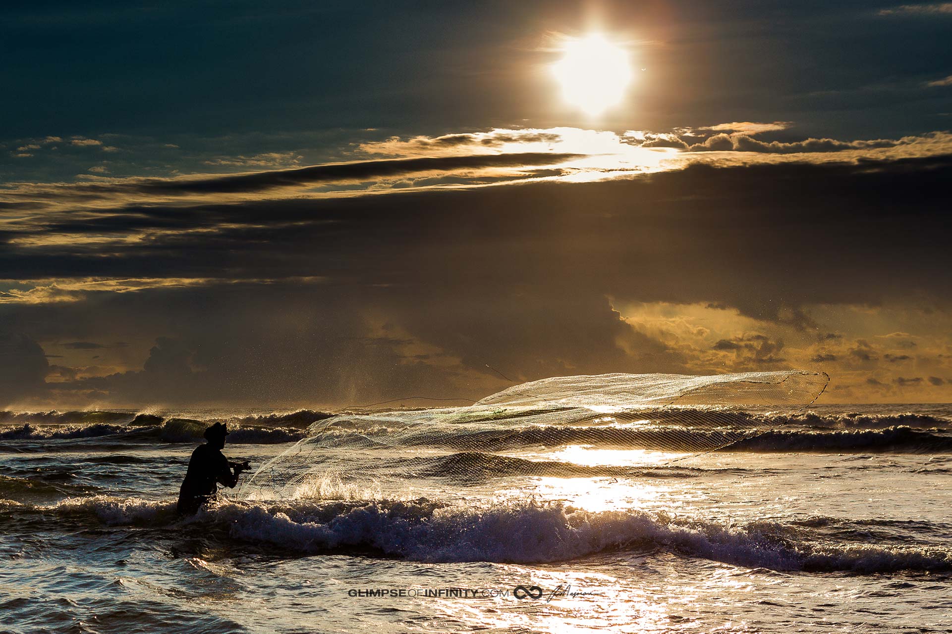 A fisherman casts out a net in the ocean under partly cloudy skies.